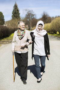 Full length of senior woman with female home caregiver walking arm in arm on street