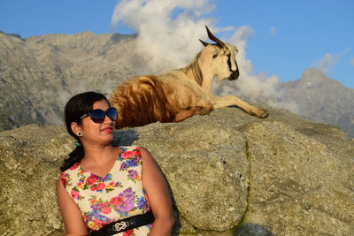 Woman in sunglasses with goat on rock against sky