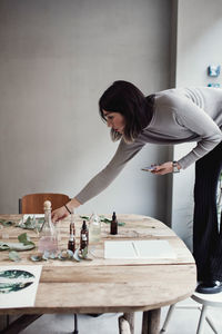 Full length of young woman standing on chair arranging table at perfume workshop