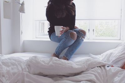 Low section of woman sitting with coffee mug on window sill in bedroom at home