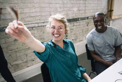 Portrait of cheerful businesswoman holding feather sitting by male colleague at desk in creative office