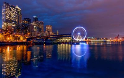 Ferris wheel by river in illuminated city against cloudy sky at night