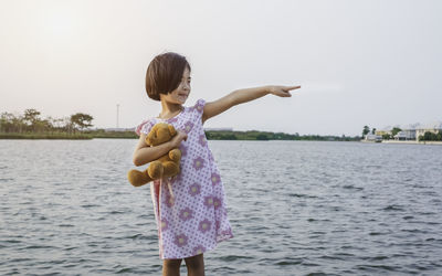 Girl pointing while holding teddy bear against lake and sky