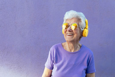 Cheerful senior female in t shirt and modern sunglasses listening to song from wireless headset on purple background