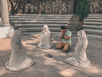 Man covering face while kneeling amidst female statues against steps