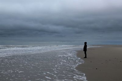 Side view of man standing on shore at beach against cloudy sky