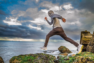 Full length of man running on moss covered rock by sea against cloudy sky