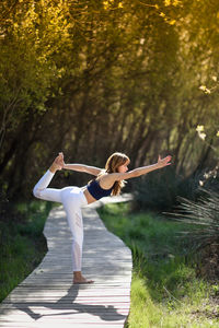 Full length of woman doing yoga on boardwalk against plants