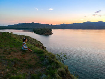 Young woman sitting on lake against sky during sunset