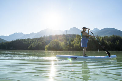 Side view of pregnant female in life vest riding sup board on calm lake against mountain ridge and forest on sunny day in british columbia, canada