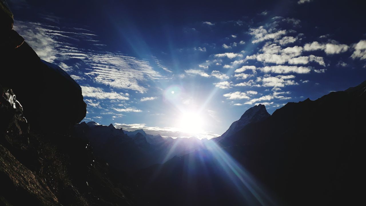 LOW ANGLE VIEW OF SUNLIGHT STREAMING THROUGH CLOUDS OVER SILHOUETTE MOUNTAIN