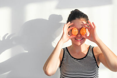 Portrait of woman holding eggs on eyes while standing against wall