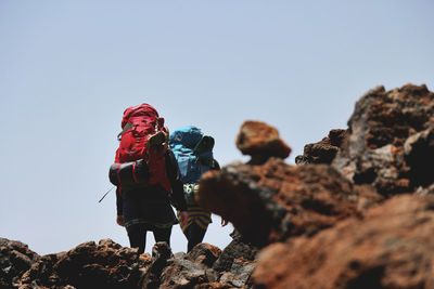 Rear view of friends with backpacks walking on rock formations against clear sky during sunny day