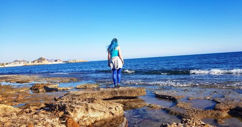 Rear view of woman standing on rock at beach against blue sky