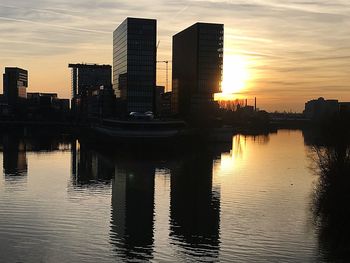 Modern buildings by river against sky during sunset