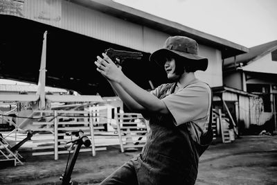 Side view of young woman holding camera while standing against built structure