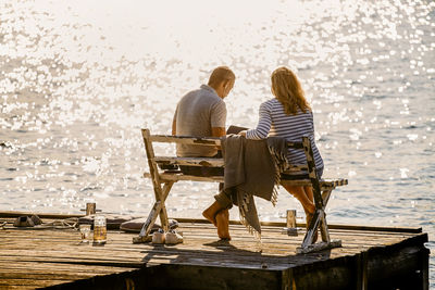 Full length of couple using laptop while sitting on bench over wooden pier against lake during summer vacation