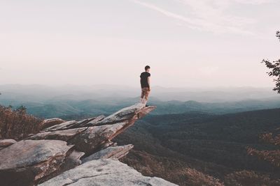 Man standing on cliff while looking at mountains against sky