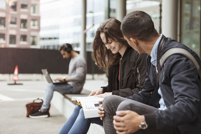 Man talking to smiling friend with book while sitting at university