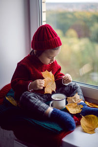 Boy a child in a red sweater and a knitted hat sits at the window on the windowsill