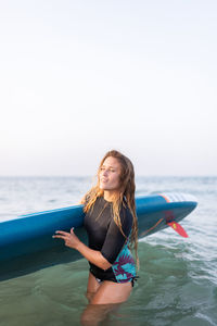 Female in swimsuit standing with sup board in sea water in summer and looking away