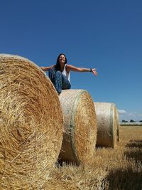 Low angle view of cheerful young woman sitting on hay bale against clear sky