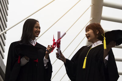 Cheerful young women holding degree certificates standing against sky