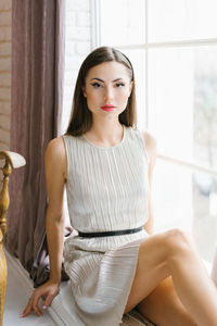 Cute stylish young woman in a festive dress with professional makeup is sitting on the windowsill