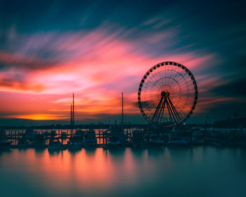 Silhouette ferris wheel by sea against sky at sunset