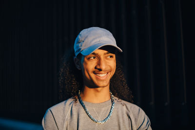 Close-up of smiling young man wearing cap looking away in dark