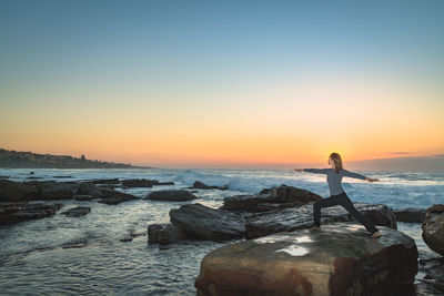 Woman exercising yoga on rock by sea against sky during sunrise