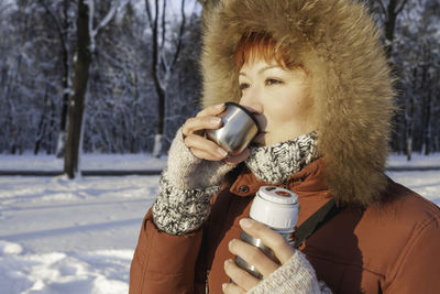 Woman in red coat drinks hot tea from metal thermos. leisure activity in winter forest at sunset.