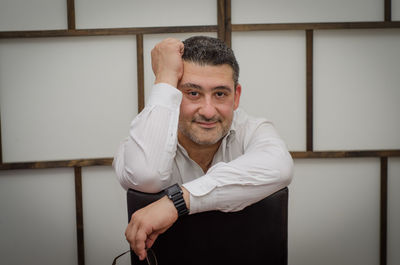 Portrait of confident man sitting on chair against wall at home