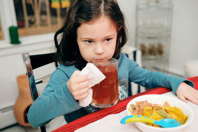 Young girl drinking herbal tea while also eating colorful bow pasta at the dinner table