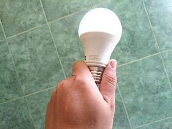 Cropped hand of man holding light bulb against tiled floor