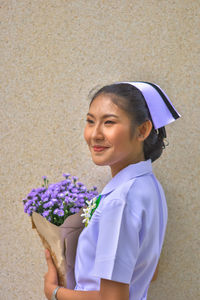 Smiling young nurse with bouquet standing against wall