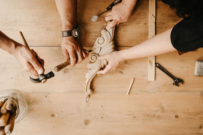 Top view of crop anonymous artisan masters with hammer and chisel carving decorative wooden detail while working together in workshop