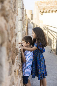 Side view of siblings looking through old window while standing outdoors