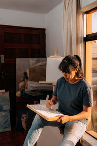 Woman at home relaxed drawing and painting close to the window at sunset