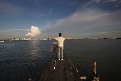 Rear view of man with arms outstretched standing on pier by sea