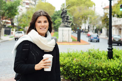 Portrait of smiling young woman holding disposable cup against plants