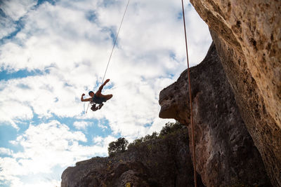 Low angle view of man hanging from safety harness by rock formation against sky