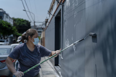 Side view of professional female painter in protective mask standing on sidewalk and painting wall using roller on city street