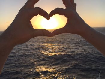 Hand holding heart shape against sea during sunset