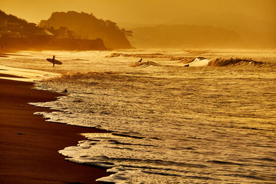 Scenic view of surfer beach with silhouette people standing against orange sky during sunrise