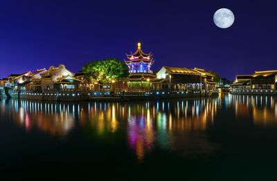 Illuminated buildings by lake with reflection against clear sky at night