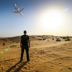 Full length rear view of man standing on desert by car against airplane flying in clear sky