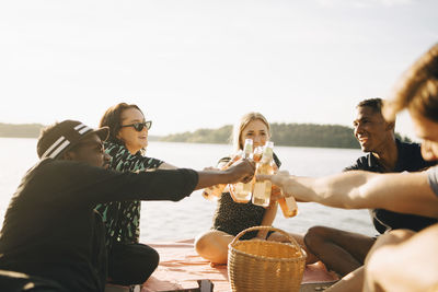 Friends toasting bottles while sitting on jetty against sky in summer