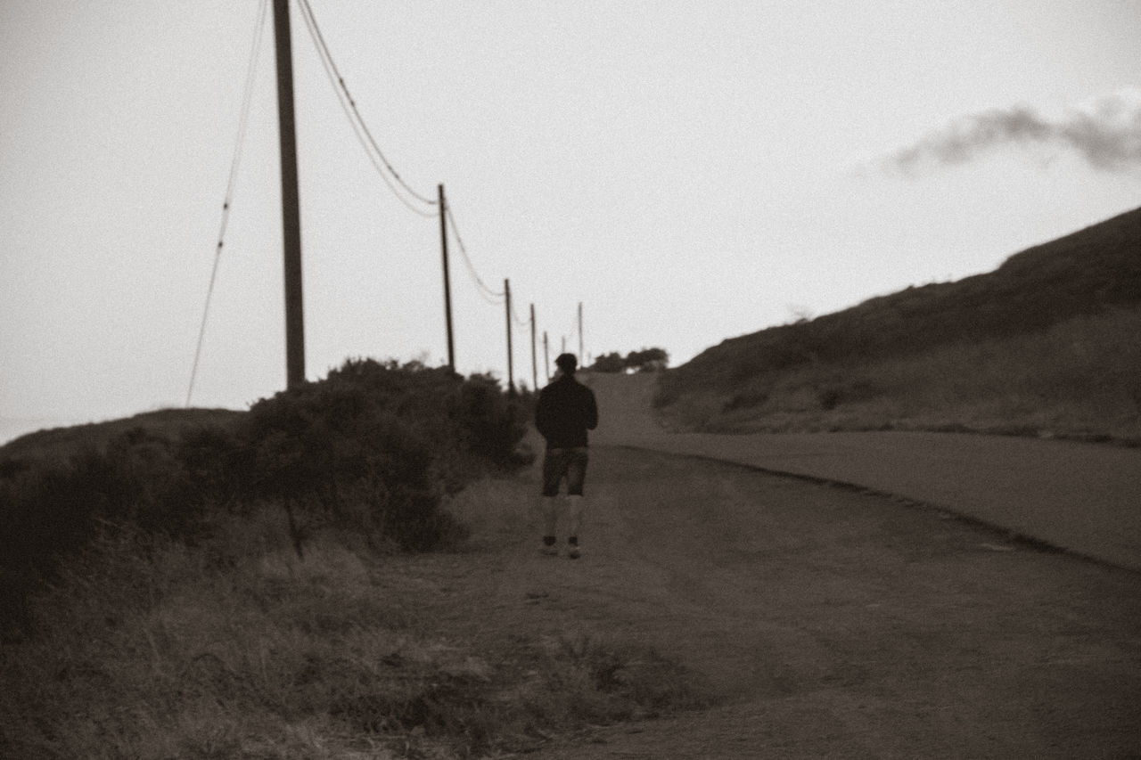 REAR VIEW OF MAN WALKING ON DIRT ROAD