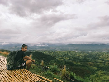 Young man sitting on mountain against sky